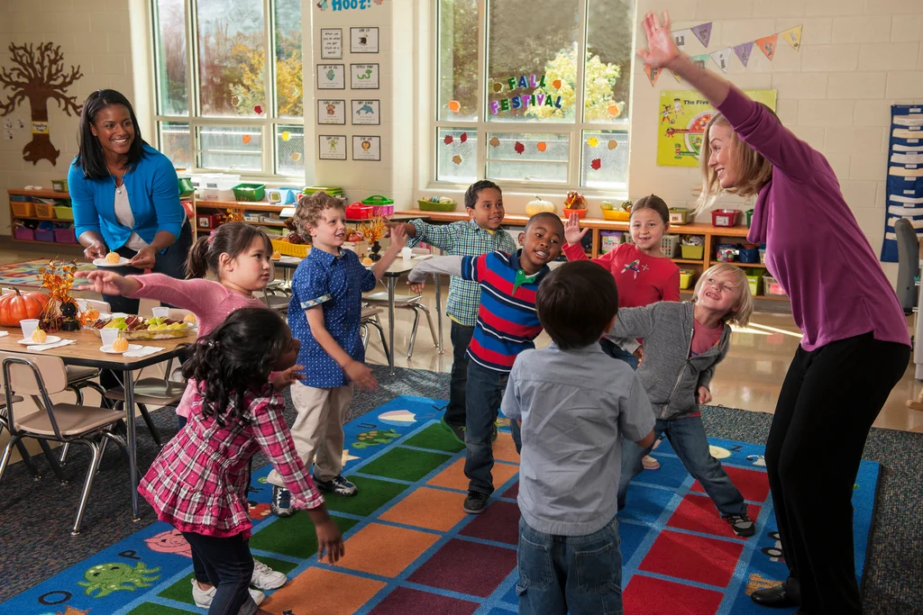 Teachers and children stretching their arms during an activity in a classroom with windows, tables and chairs