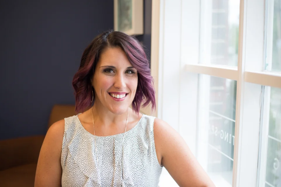 A smiling person with short purple hair and a patterned blouse seated beside a window